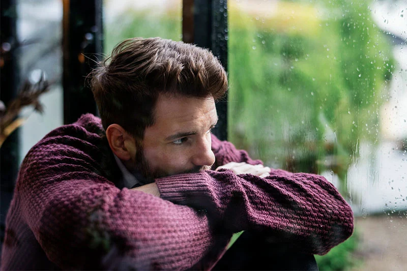 Depressed man looking out the window thinking about getting treatment for substance use disorder in New Jersey. The importance of an integrated approach to drug addiction treatment emphasizes the need to address mental health issues alongside addiction recovery